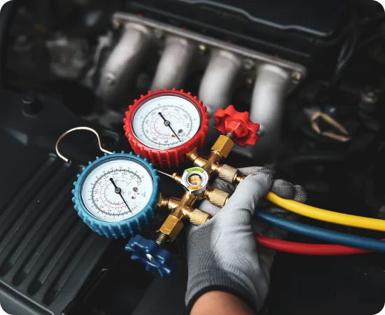 A gloved hand holds a manifold gauge set with blue, red, and yellow hoses in front of a car engine, used for checking automotive air conditioning pressure.