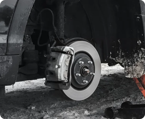 Close-up of a cars exposed front wheel hub and brake disc, with the wheel removed. The car is parked on a snowy, icy surface, and the wheel well is partially covered in mud and snow.