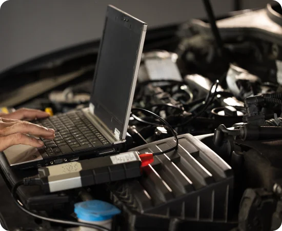 A person uses a laptop connected to a diagnostic tool under the hood of a car, likely for vehicle diagnostics or troubleshooting engine issues.
