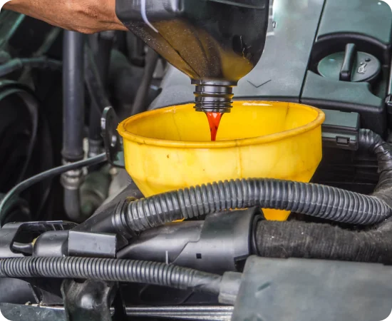A person pours red engine oil from a black container into a yellow funnel placed in a car engine bay. Various engine components and hoses are visible around the funnel.