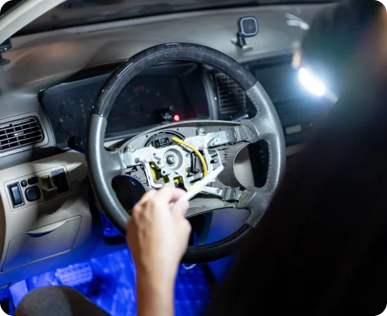 A person is working on a car’s steering wheel with the airbag removed, exposing wires and internal components. The dashboard and interior are dimly lit, and a bright light illuminates the work area.