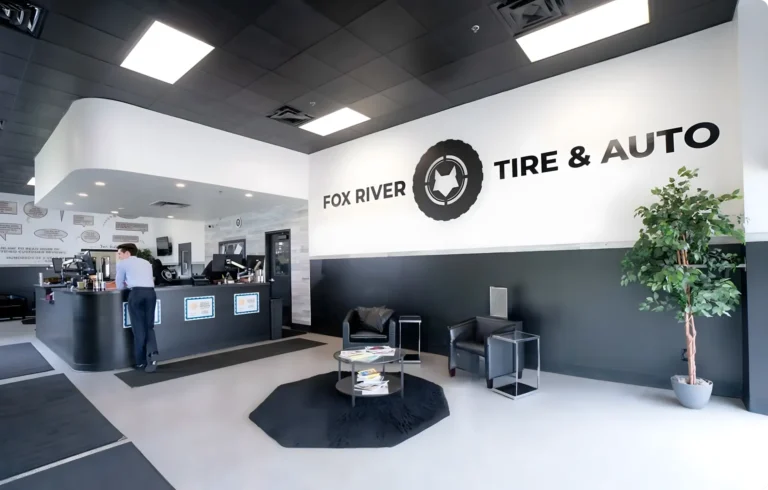 Modern auto shop waiting area with black and white decor, a large Fox River Tire & Auto sign on the wall, two black chairs, a table with magazines, a potted plant, and a counter where a person is assisting a customer.