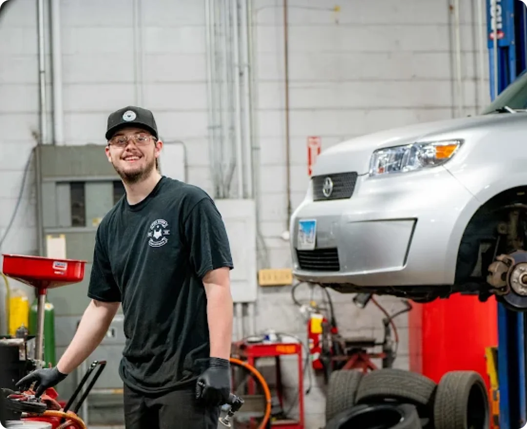 A mechanic wearing glasses, a black cap, black t-shirt, and gloves smiles while standing in an auto repair shop near equipment. A silver car is lifted on a hydraulic lift in the background.