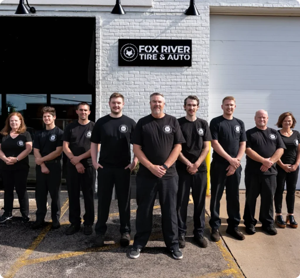 Nine people in matching black shirts stand in front of a white building with a Fox River Tire & Auto sign, posing for a group photo on a paved lot.