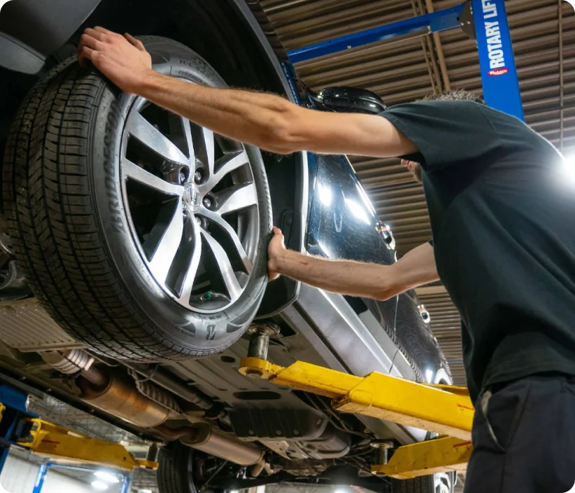 A mechanic inspects a car’s front tire on a hydraulic lift in an auto repair shop, checking for issues or maintenance needs.