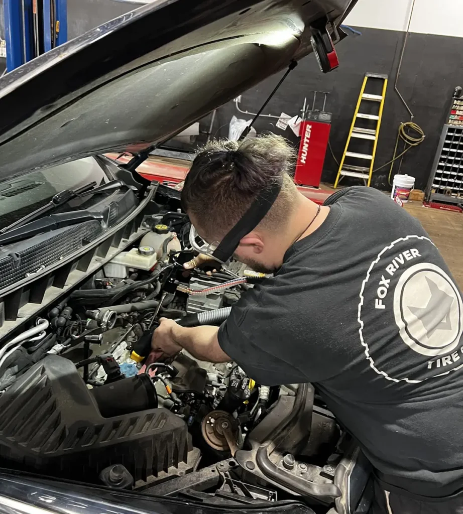 A mechanic wearing a Fox River Tire shirt works under the hood of a car in a garage with tools, equipment, and a yellow ladder in the background.