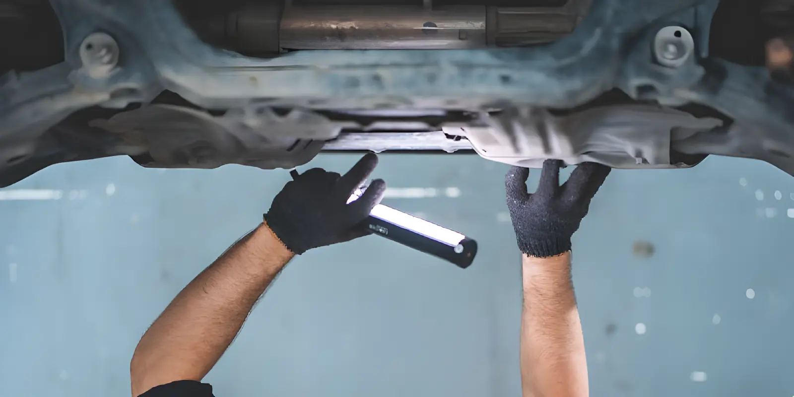 A person wearing black gloves inspects the underside of a vehicle with a handheld flashlight, illuminating car components in a workshop setting.