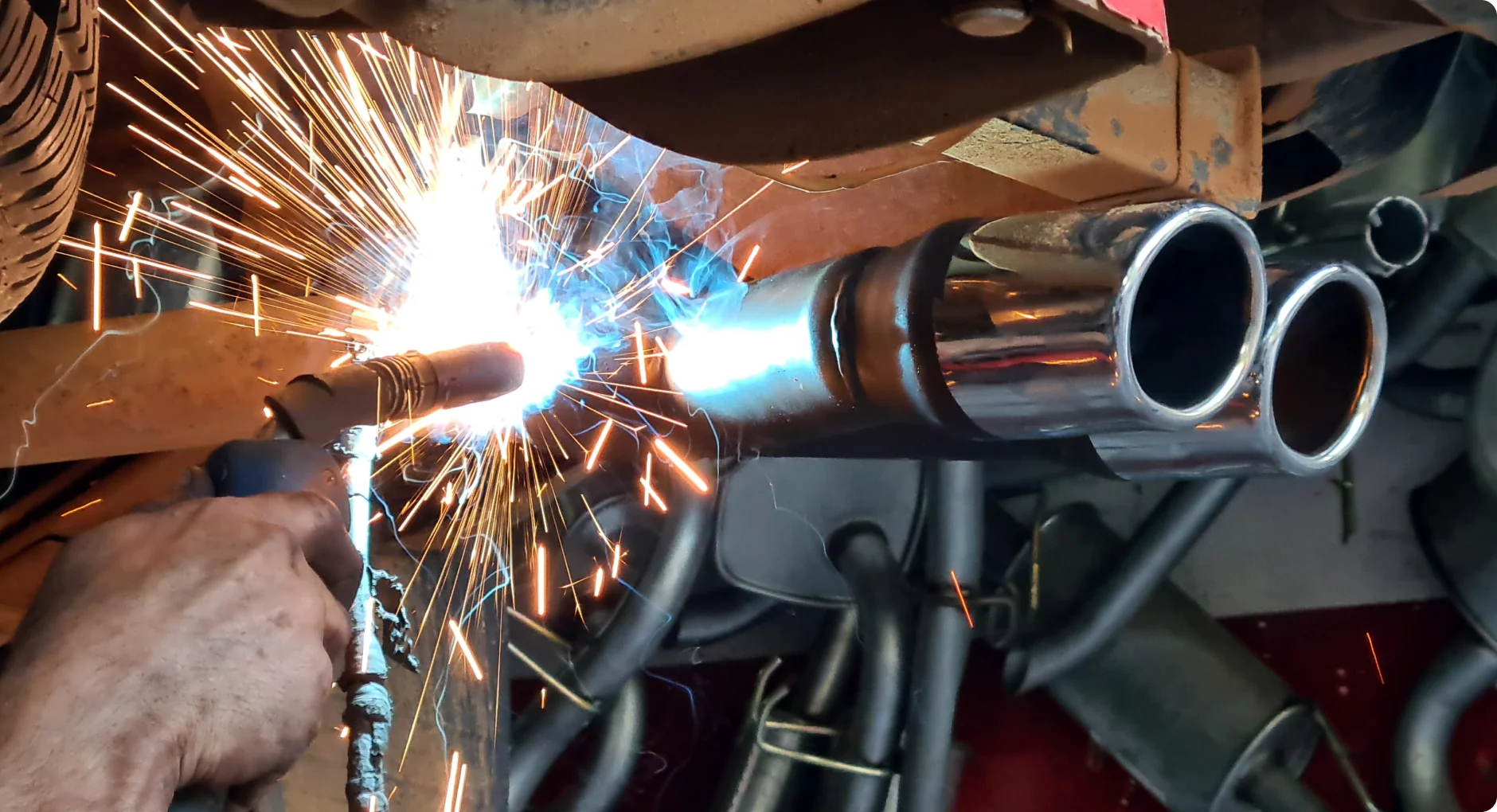 A person uses a welding torch on a metal exhaust pipe, producing bright sparks and a blue-white light. The surrounding area contains various metal parts and tubes.