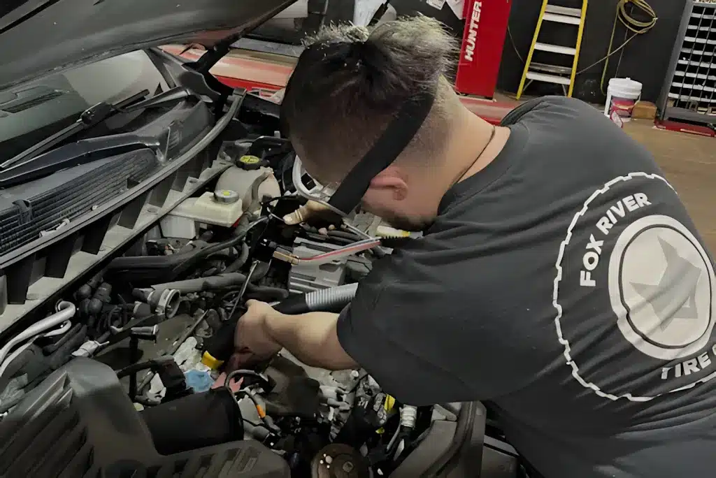 A mechanic wearing a Fox River Tire shirt works on a car engine in an auto repair shop, with the vehicle’s hood open and various tools and equipment visible in the background.