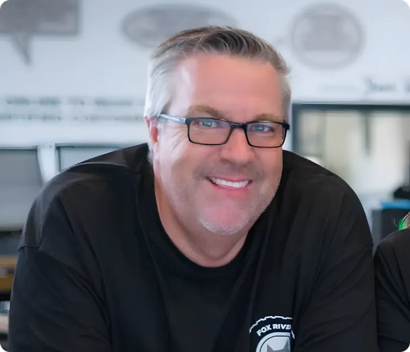 A smiling man with short gray hair and glasses wears a black shirt with a white logo. He is indoors, in an office or workspace setting, with a blurred background, reflecting the welcoming spirit of our about us story.