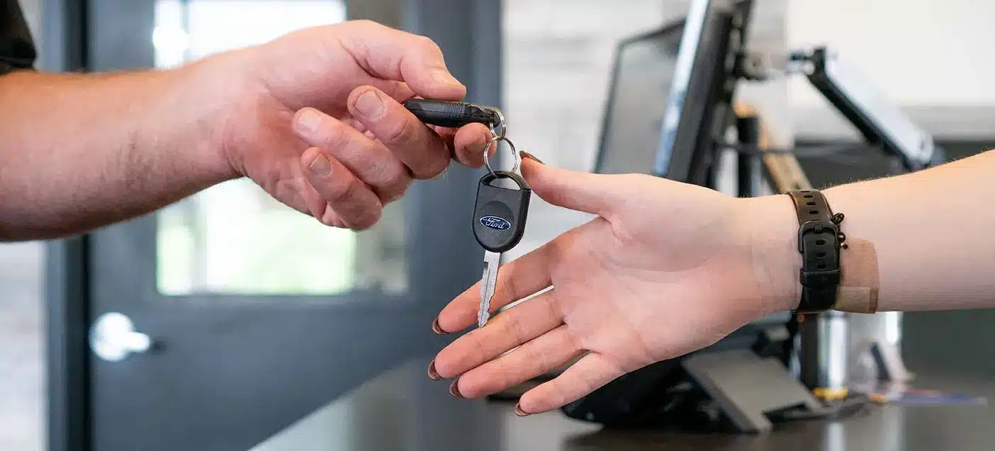 A person hands over a car key to another person indoors, with a computer monitor and door visible in the background.