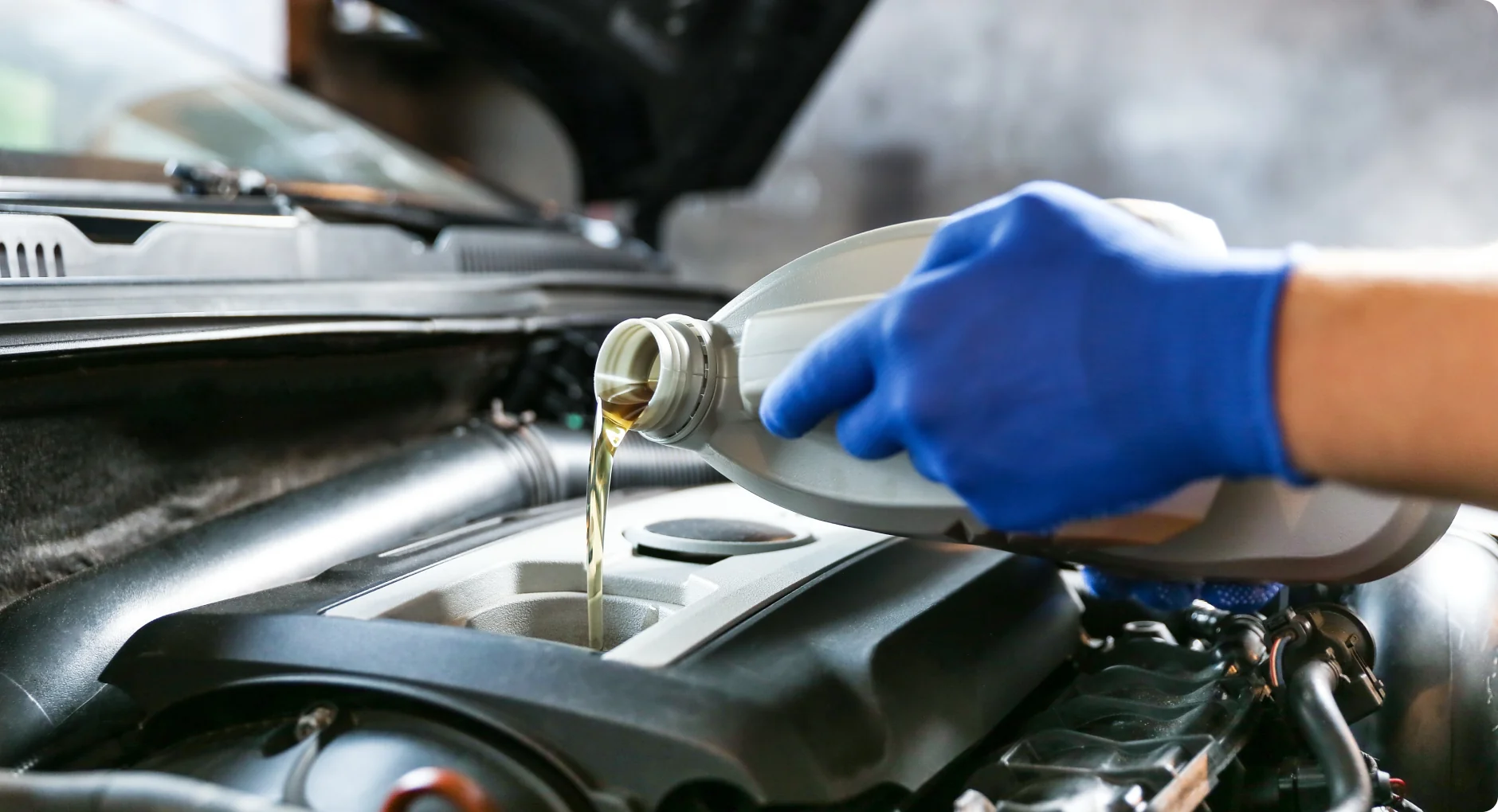 A person wearing blue gloves pours motor oil from a plastic bottle into a car engine, with the car hood open in the background.