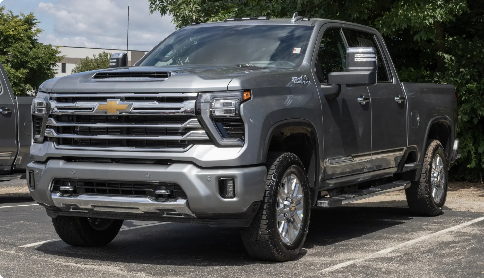 A gray Chevrolet Silverado HD pickup truck is parked in a lot near green trees, with a clear sky and some buildings in the background.
