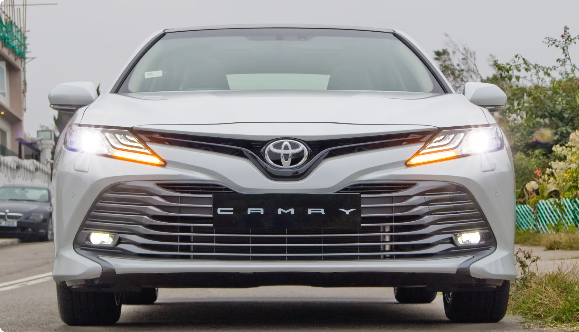 Front view of a silver Toyota Camry with headlights and fog lights on, parked on a street. The Toyota emblem is centered above the grille, and CAMRY is displayed on the license plate area.