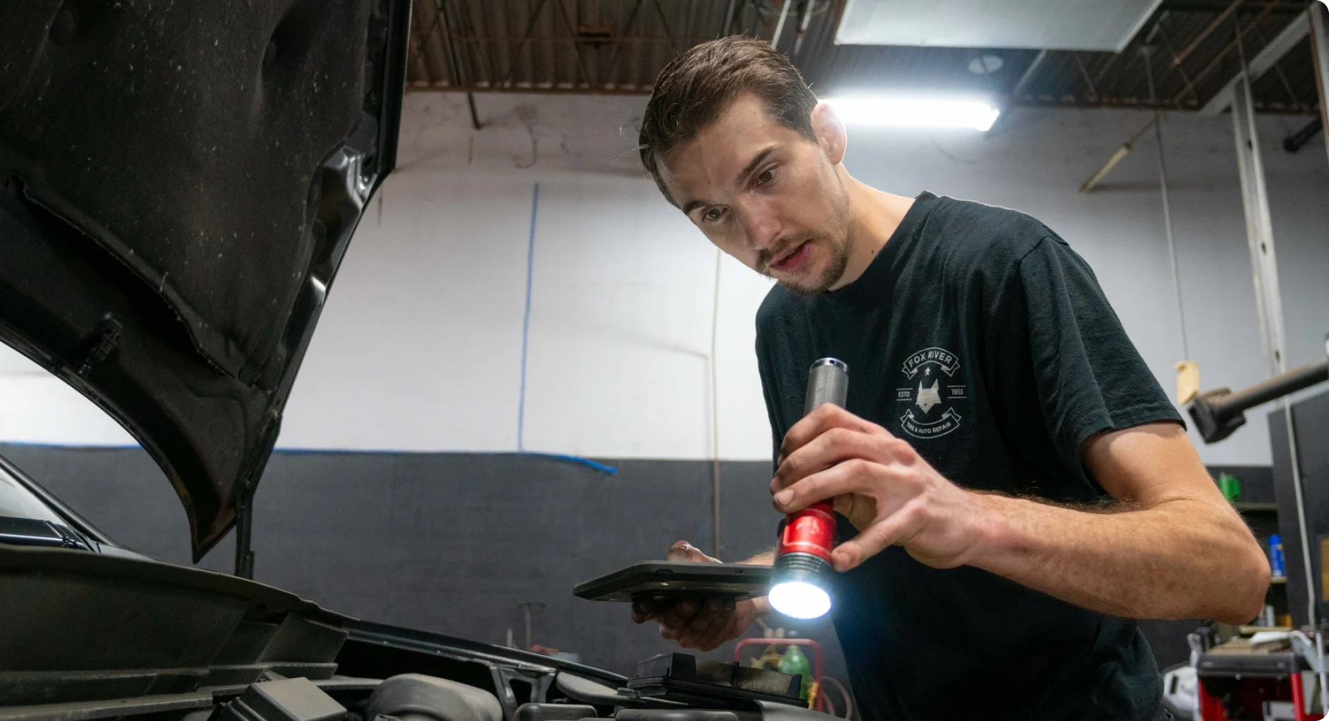 A mechanic inspects a car engine, holding a flashlight in one hand and a tablet in the other, under the open hood in a garage with exposed ceiling beams.