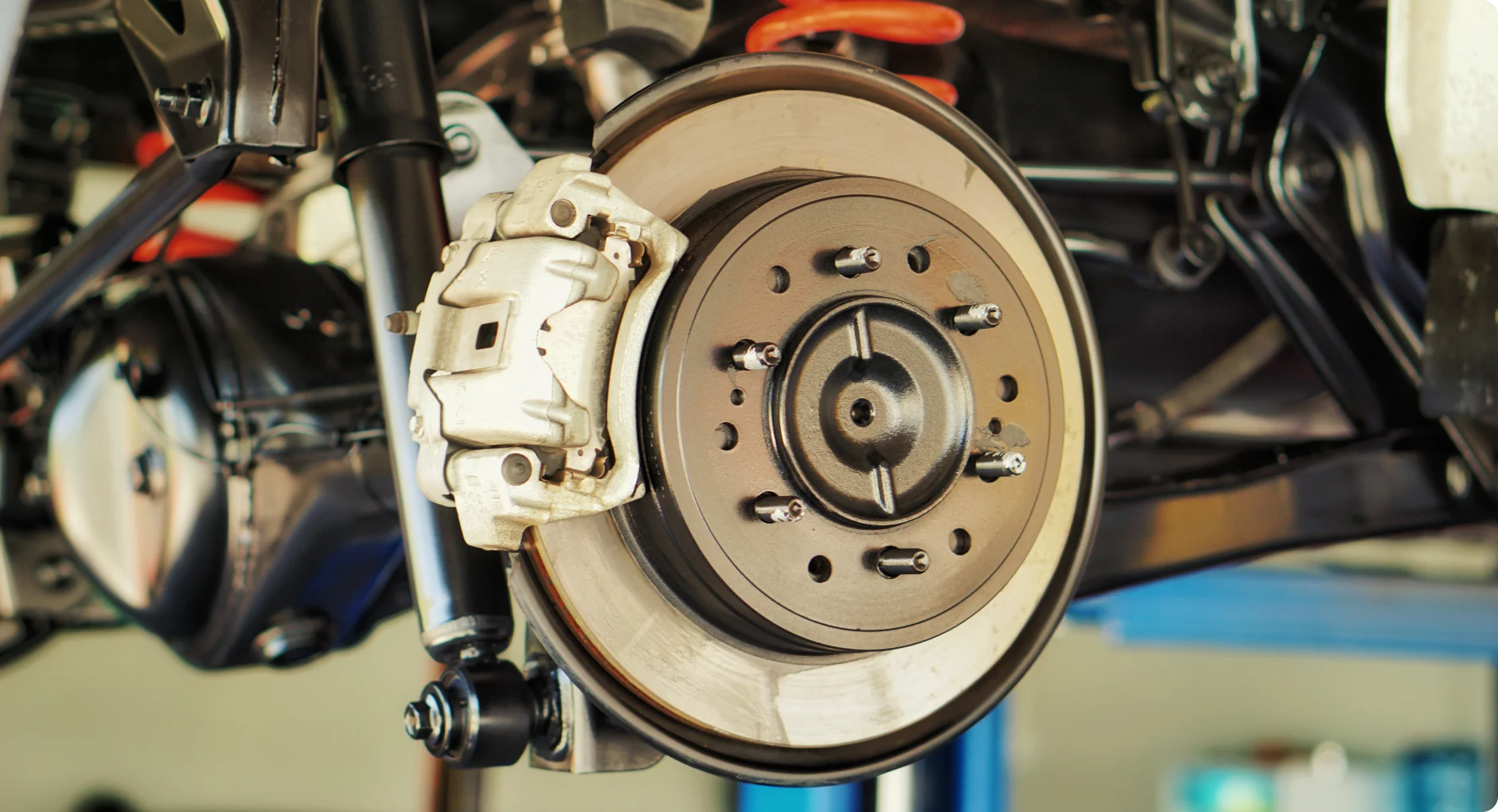 Close-up view of a car’s disc brake system, showing the brake rotor, caliper, and suspension components, with the car lifted off the ground in a workshop setting.