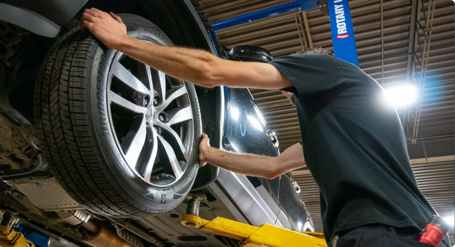 A mechanic installs or removes a tire from a car that is lifted on a yellow hydraulic lift in an auto repair shop. The mechanic is wearing a black shirt and is working under bright overhead lights.