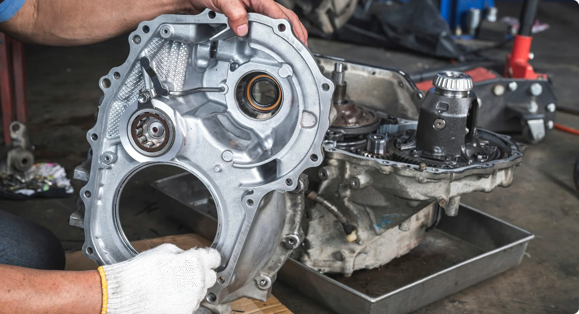A person wearing a white glove holds a metal transmission casing next to an exposed car transmission with visible gears and parts in a workshop setting.