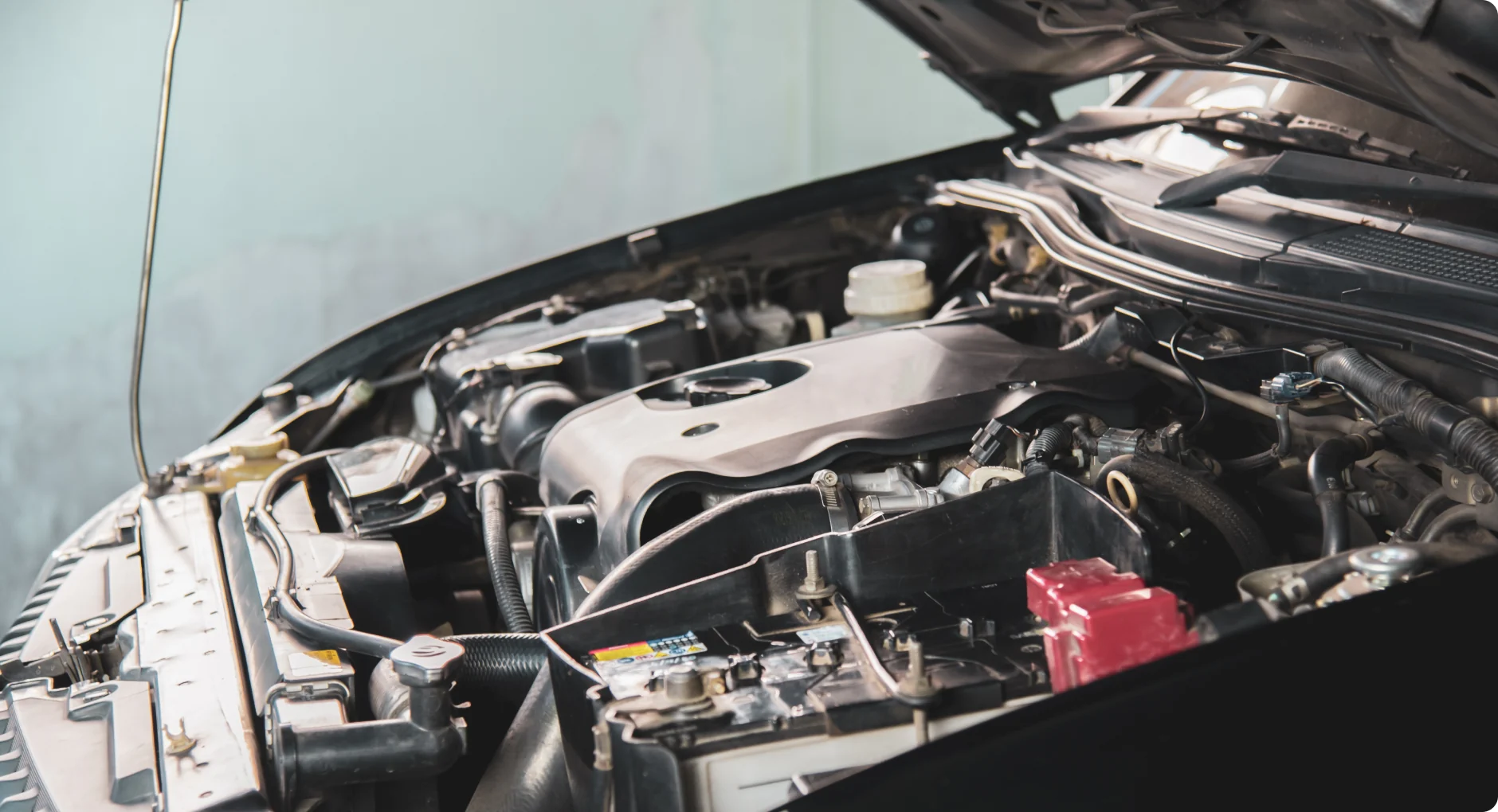 Close-up view of a car engine with the hood open, showing various engine components including the battery, hoses, and engine cover. The background is out of focus, highlighting the engine area.