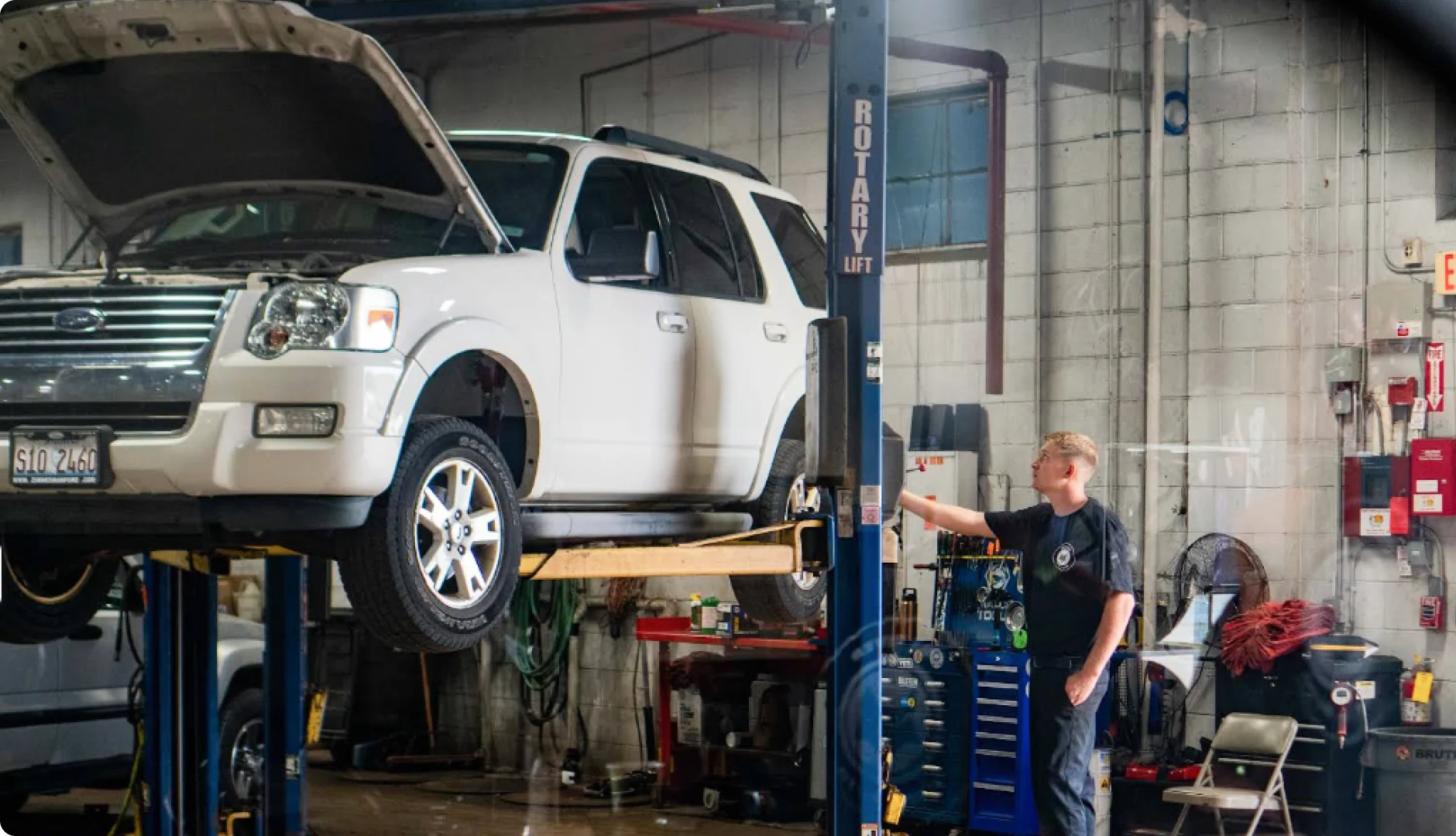 A white SUV with its hood open is elevated on a car lift in an auto repair shop. A mechanic stands nearby, operating the lift controls. The shop contains various tools and equipment.