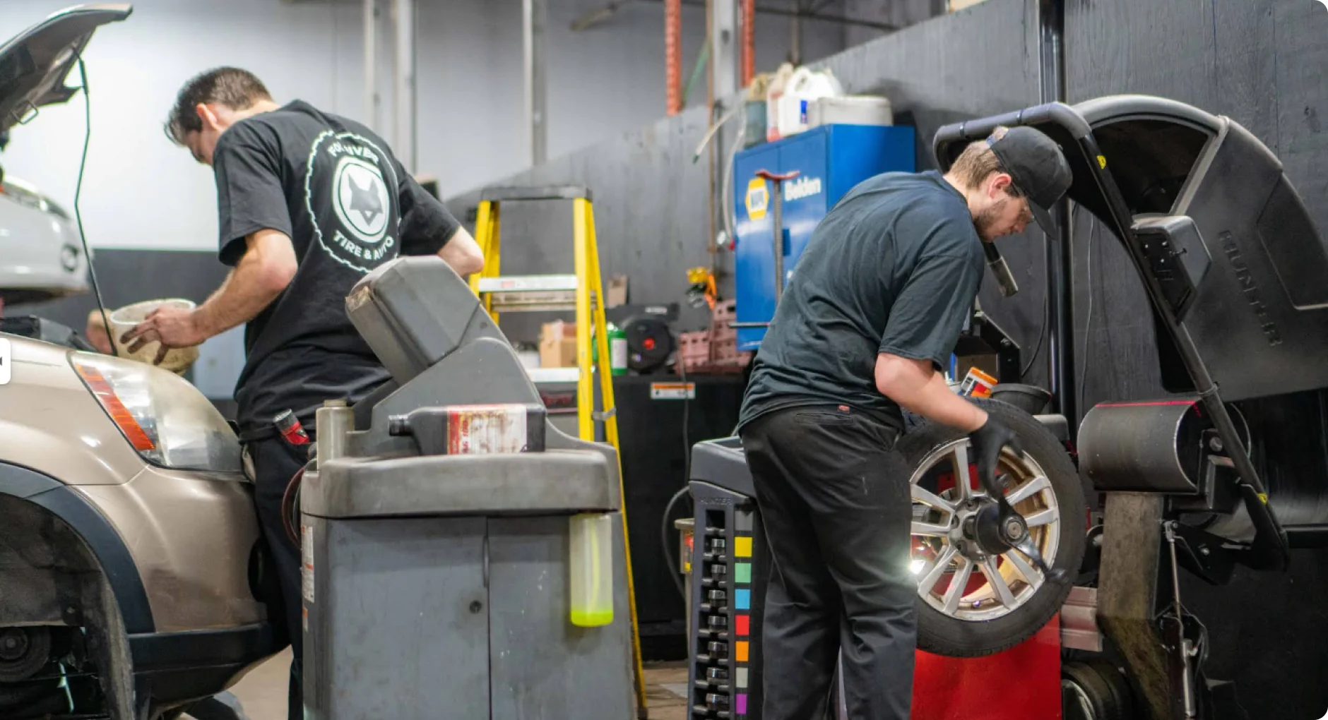 Two auto mechanics work in a garage; one is repairing a car on the left while the other uses a tire balancing machine on a wheel in the foreground. Various tools and equipment are visible around them.