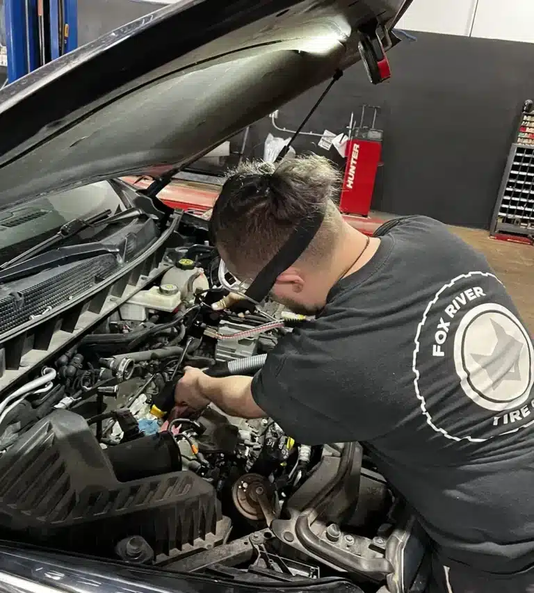 A mechanic wearing a Fox River Tires shirt works under the hood of a car in an auto repair garage, focusing on the engine. Tools and equipment are visible in the background.