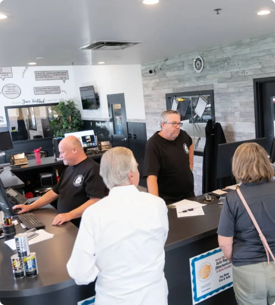 Two men in black shirts assist two customers at a modern, circular front desk. One works at a computer while the other speaks with customers. The background includes an about us bulletin board, gray wall, and framed documents.